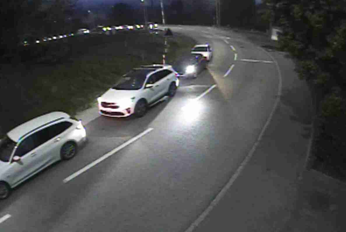 Nachtaufnahme einer Überwachungskamera einer kurvigen Landstrasse mit Mittellinie. Zwei weisse Personenwagen fahren hintereinander in eine Richtung, ein weiteres Fahrzeug mit eingeschaltetem Fernlicht kommt entgegen. Fahrbahn ist trocken, im Hintergrund weitere Fahrzeuglichter erkennbar. Strasse ist beidseitig von Vegetation gesäumt und macht einen kurvenreichen Verlauf.