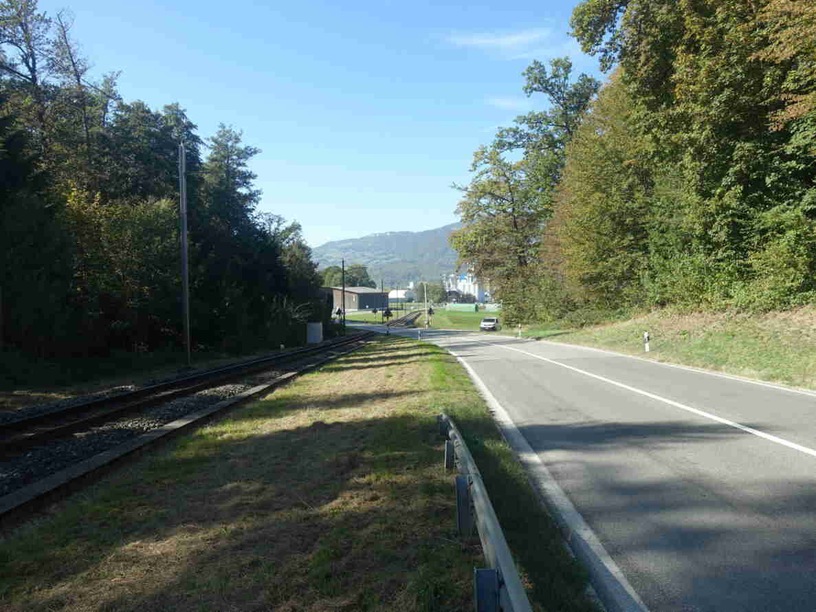 Überlandstrasse mit Mittellinie und Leitplanke, parallel zu einer eingleisigen Bahnstrecke geführt. Am Horizont ein Bahnübergang mit Andreaskreuzen und Warnsignalen, wo die Strasse die Gleise kreuzt. Ein Kleintransporter fährt auf der Strasse Richtung Bahnübergang. Der Asphalt ist trocken und in gutem Zustand. Im Hintergrund Hügelzüge sowie ein Gebäude mit Silo.