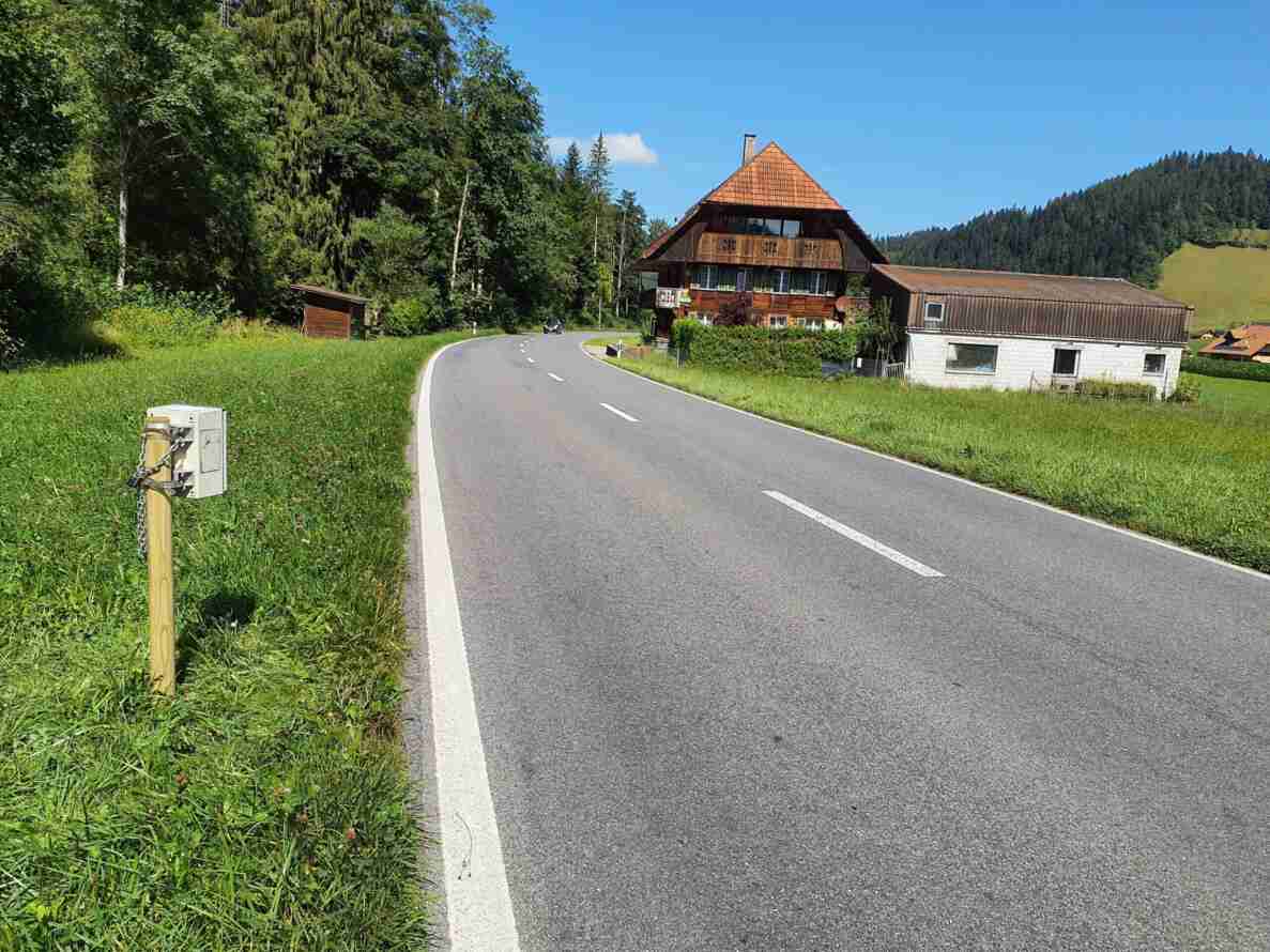 Kurvige, zweispurige Überlandstrasse mit Mittellinie in gutem Zustand, am rechten Fahrbahnrand ein Holzpfosten mit montiertem Seitenradargerät zur Geschwindigkeitsmessung. Ein Personenwagen nähert sich aus der Ferne. Im Hintergrund ein traditionelles Bauernhaus mit Schindelfassade und Nebengebäude, umgeben von Wiesen, Wald und bewaldeten Hügeln.