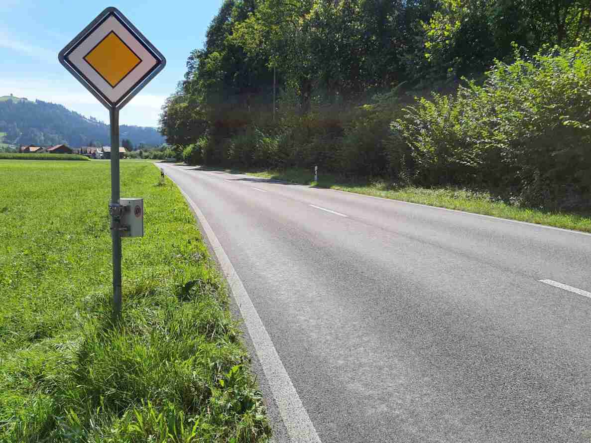 Hauptstrasse ausserorts mit Signal «Hauptstrasse» am Strassenrand, an dessen Mast ein seitlich montiertes Messgerät zur Verkehrsmessung angebracht ist. Die Fahrbahn ist in gutem Zustand, führt in einer Kurve durch grüne Wiesen und Wald, im Hintergrund Hügel und einzelne Gebäude. Keine Fahrzeuge oder Personen sichtbar.