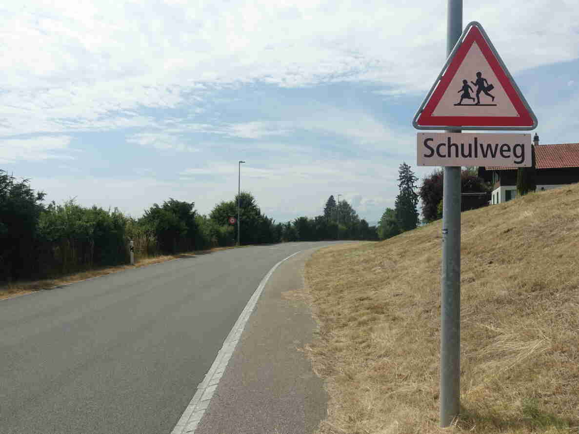 Warntafel «Kinder» mit Zusatztafel «Schulweg» am Strassenrand der Dorfstrasse in Alchenstorf. Die schmale, leicht kurvige Überlandstrasse verläuft ohne Trottoir zwischen Hecken und einer trockenen Böschung, im Hintergrund eine Tempo-50-Tafel sowie Strassenlaternen. Keine Verkehrsteilnehmenden sichtbar, die Fahrbahn ist frei.
