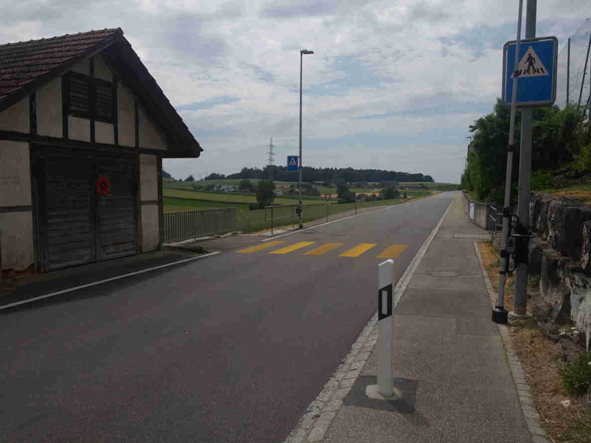 Dorfstrasse in Alchenstorf mit gelb markiertem Fussgängerstreifen und Fussgängerstreifen-Signal am rechten Strassenrand. Links ein altes Bauernhaus mit Fachwerk und Parkverbotsschild, rechts ein Trottoir entlang einer Steinmauer. Die zweispurige Strasse führt geradeaus durch offenes Landwirtschaftsgebiet, im Hintergrund ist ein weiteres Fussgängerstreifen-Signal sowie ein Vortrittssignal sichtbar. Kein Verkehr auf der Strasse erkennbar.