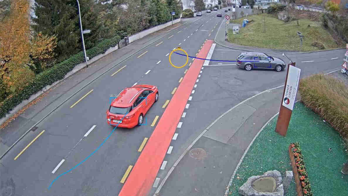 Vogelperspektive einer mehrspurigen Strasse mit rot markiertem Radstreifen und Tramgleisen. Ein rotes und ein graues Auto sind mit farbigen Trajektorien und Geschwindigkeitswerten zur Verkehrsmessung markiert. Rechts zweigt eine Nebenstrasse ab, Verkehrsschilder regeln die Vorfahrt. Am Strassenrand stehen Vegetation und ein Wegweiser zu Ferrer Garden. Im Hintergrund weitere Fahrzeuge und Wohnbebauung.