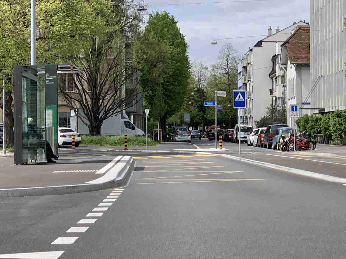 Haltestelle Wielandplatz mit moderner Glas-Wartehalle für Buslinien 33 und 48, in der eine sitzende Person wartet. Die zweispurige Strasse mit gelber Fahrbahnmarkierung ist mässig befahren, am Fahrbahnrand parkieren mehrere Autos und Velos. Ein Fussgängerstreifen wird durch ein blaues Signal angekündigt, daneben weist ein Hinweis auf einen kombinierten Fuss- und Radweg hin. Fahrbahn und Trottoirs sind neu asphaltiert.