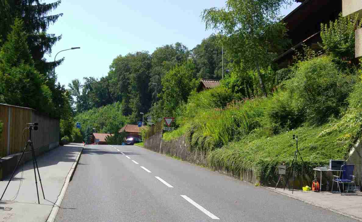 Strasse in Bern mit beidseitig aufgestellten Stativen mit Mikrofonen zur akustischen Messung. Rechts ein Klapptisch mit Messgeräten und ein Stuhl. Die Fahrbahn ist frei von Verkehr, ein Auto steht am Strassenrand. Verkehrsschilder zeigen ein Fussgängerstreifen-Signal sowie ein Vortrittssignal mit Zusatztafel «50 m». Die Strasse ist asphaltiert und in gutem Zustand, gesäumt von Bäumen und einer Steinmauer.