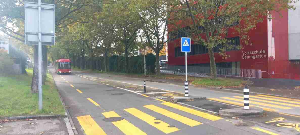 Strasse mit gelb markiertem Fussgängerstreifen vor der Volksschule Baumgarten, gekennzeichnet durch ein Fussgängerstreifen-Signal und schwarz-weiss gestreifte Leitpfosten. Ein roter Linienbus nähert sich auf der Fahrbahn. Am linken Bildrand steht eine Verkehrstafel mit einer Messeinrichtung. Der Strassenbelag ist trocken und mit Laub bedeckt, die Fahrbahnmarkierungen sind gut sichtbar.