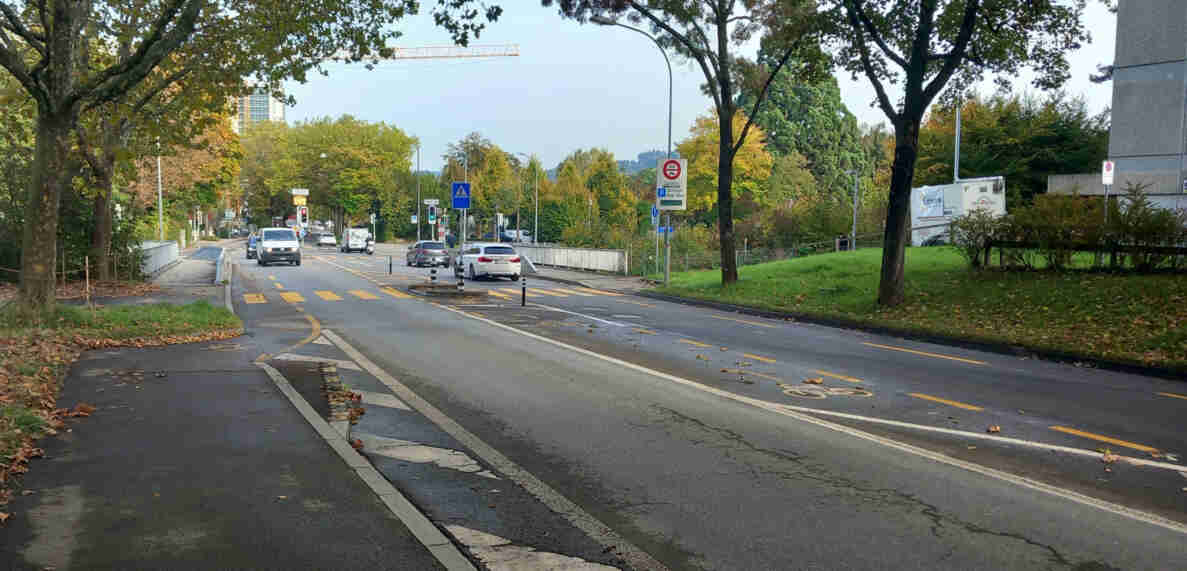 Ostermundigenstrasse mit neu markiertem gelbem Fussgängerstreifen, flankiert von Radstreifen mit Velosymbolen. Ein blaues Signal weist auf den Fussgängerstreifen hin, ein Hinweisschild zeigt Richtung Zentrum Paul Klee. Die Ampel für die Geradeausspur zeigt Grün, mehrere Autos fahren auf der leicht rissigen Fahrbahn Richtung Stadt. Rechts Grünfläche mit Bäumen, im Hintergrund ein Baukran und Hochhaus.