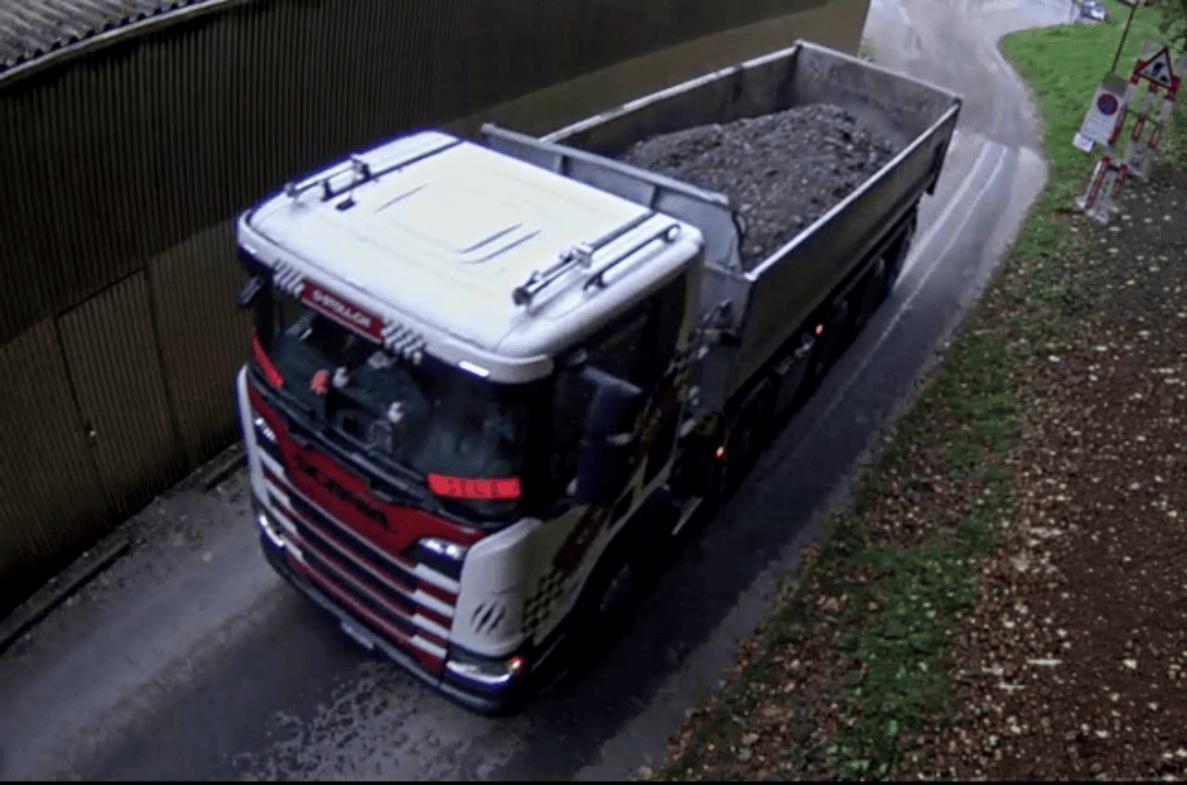 Videostandbild aus Bern-Schwellenmätteli: Kamerablick von oben auf einen mit Aushubmaterial beladenen Lastwagen, der auf einer schmalen, engen Strasse zwischen einer Holzwand und einem Grünstreifen unterwegs ist. Am rechten Strassenrand steht ein rot-weisses Absperrgitter mit Verkehrssignal. Die Fahrbahn bietet nur Platz für ein Fahrzeug, Personen sind keine sichtbar.