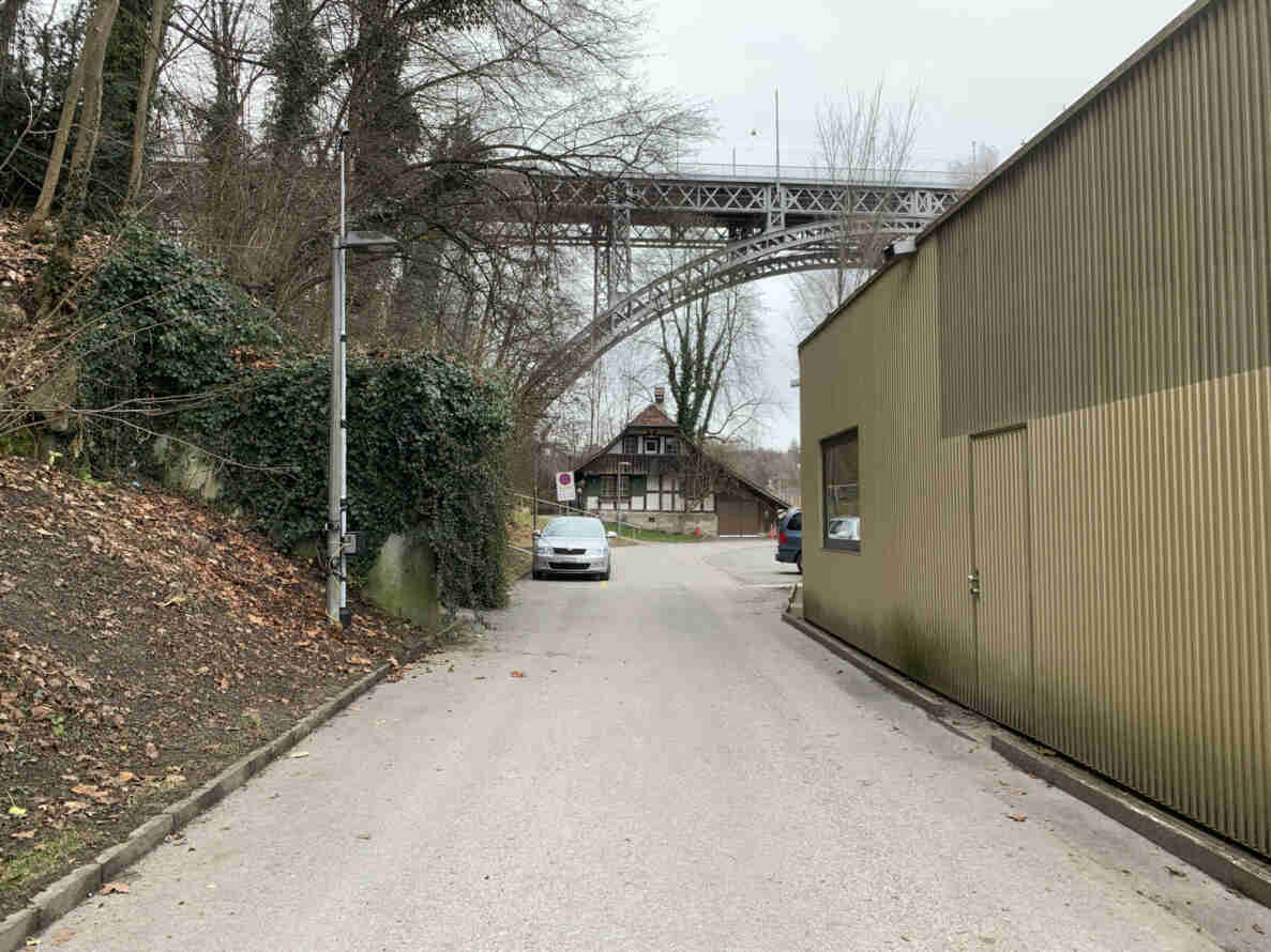 Schmale Zufahrtsstrasse mit trockenem, intaktem Belag, seitlich von Efeu-bewachsener Böschung und einer Lagerhalle gesäumt. An einem Mast ist eine installierte Langzeitkamera zur Messung montiert. Im Hintergrund führt eine stählerne Bogenbrücke über die Strasse, davor ein Fachwerkhaus. Ein Halteverbotssignal steht am Strassenrand, mehrere parkierte Autos sind sichtbar, keine Personen im Bild.