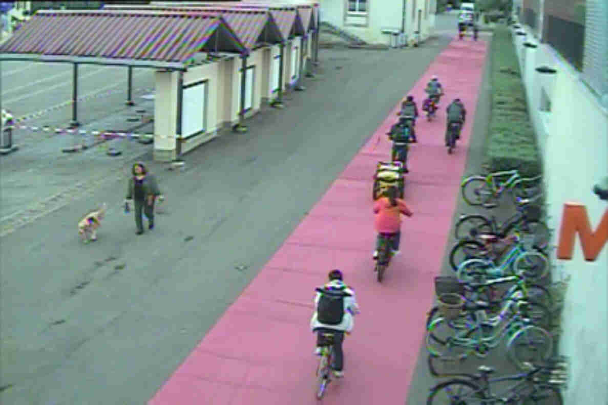 Binningen Dorfplatz: Mehrere Velofahrende nutzen den roteingefärbten Radweg entlang eines gepflasterten Platzes. Rechts stehen zahlreiche Fahrräder in Veloständern aufgereiht, links befindet sich ein überdachter Marktstand-Bereich mit gestreiftem Vordach. Eine Person mit Hund überquert den angrenzenden Fussgängerbereich. Die Markierung des Radstreifens trennt den Veloverkehr klar vom übrigen Platz.