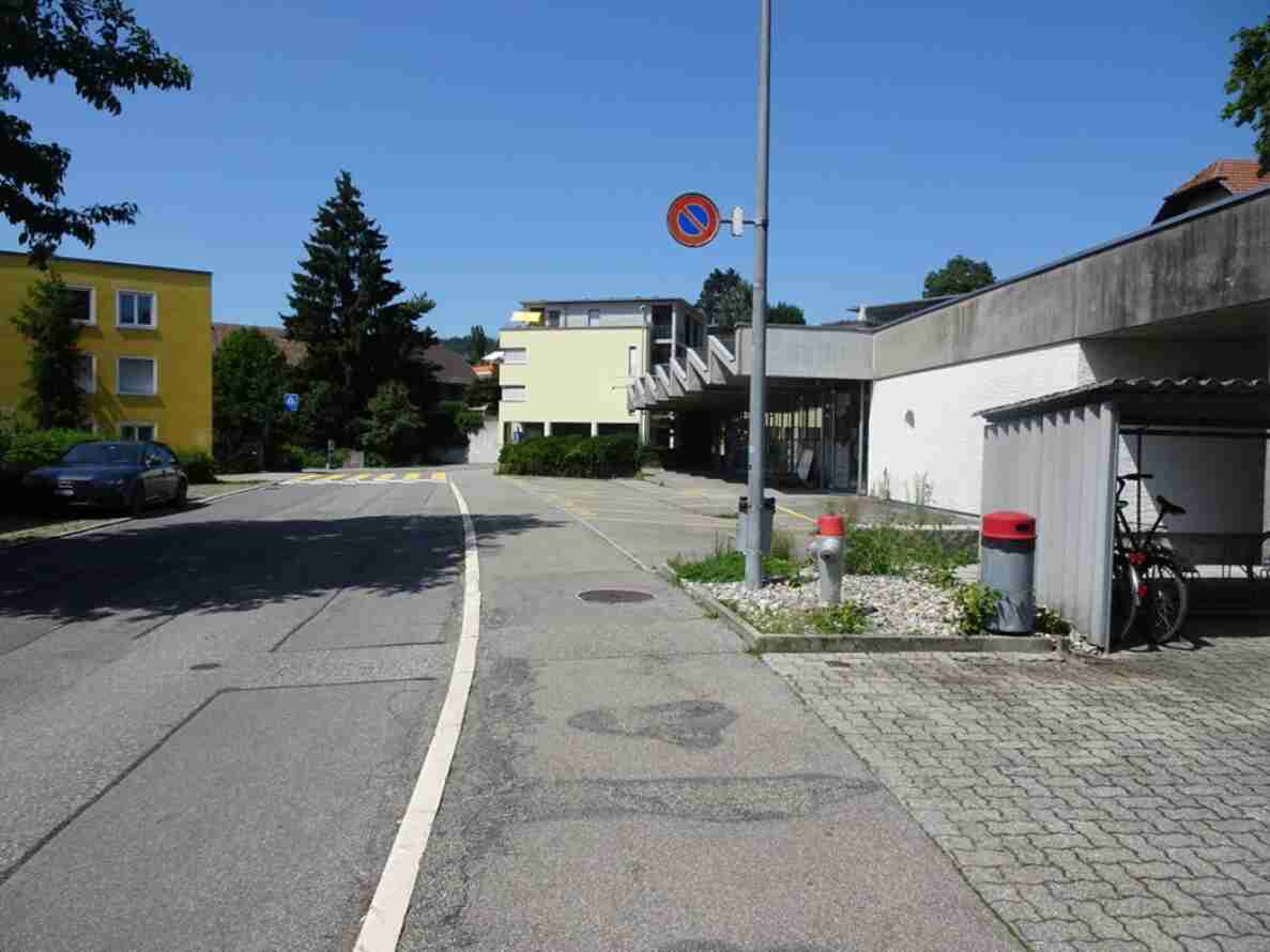Hühnerbühlstrasse in Bolligen vor Baubeginn der Wohnüberbauung, Blickrichtung Norden. Schmale Quartierstrasse mit Randstein und Trottoir, gelbe Bodenmarkierung für eine Fussgängerquerung, Halteverbotssignal an einem Mast. Parkfelder vor mehrstöckigen Wohnhäusern, ein parkiertes Auto am Strassenrand. Rechts ein überdachter Veloabstellplatz mit Fahrrad, ein Hydrant sowie ein Robidog- und ein Abfallbehälter. Keine Personen auf der Strasse sichtbar.