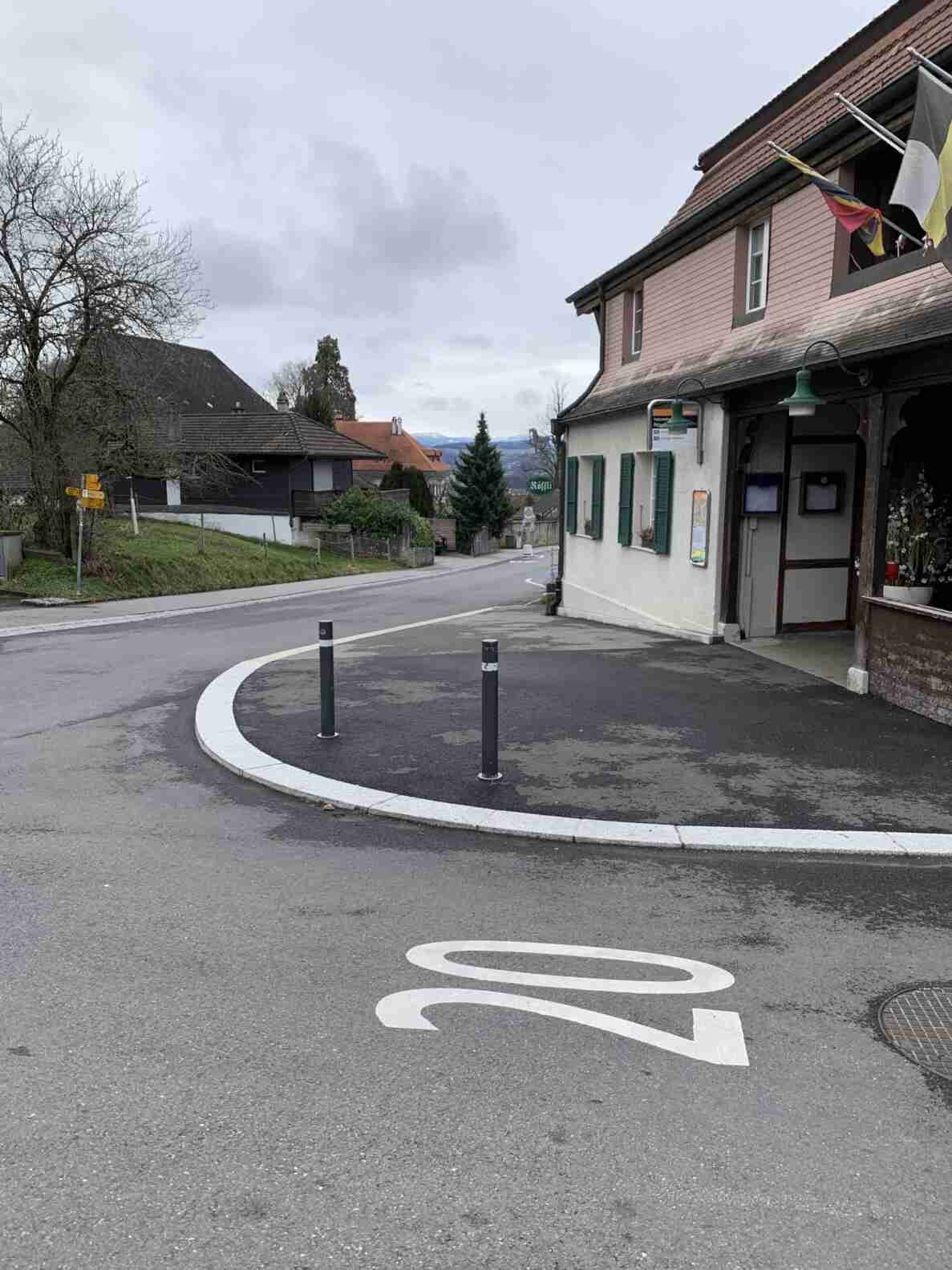 Strassenkreuzung mit Bushaltestelle beim Restaurant Rössli, gekennzeichnet durch ein Verkehrsschild und einen Fahrplankasten an der Hausfassade. Auf der nassen Fahrbahn ist eine Buslinien-Markierung «20» mit Richtungspfeil aufgemalt. Zwei schwarze Poller trennen den Vorplatz von der Strasse. Im Hintergrund weist eine Wegweiser-Tafel für Fussgänger auf verschiedene Ziele hin, die Strasse verläuft leicht bergab in eine Kurve.