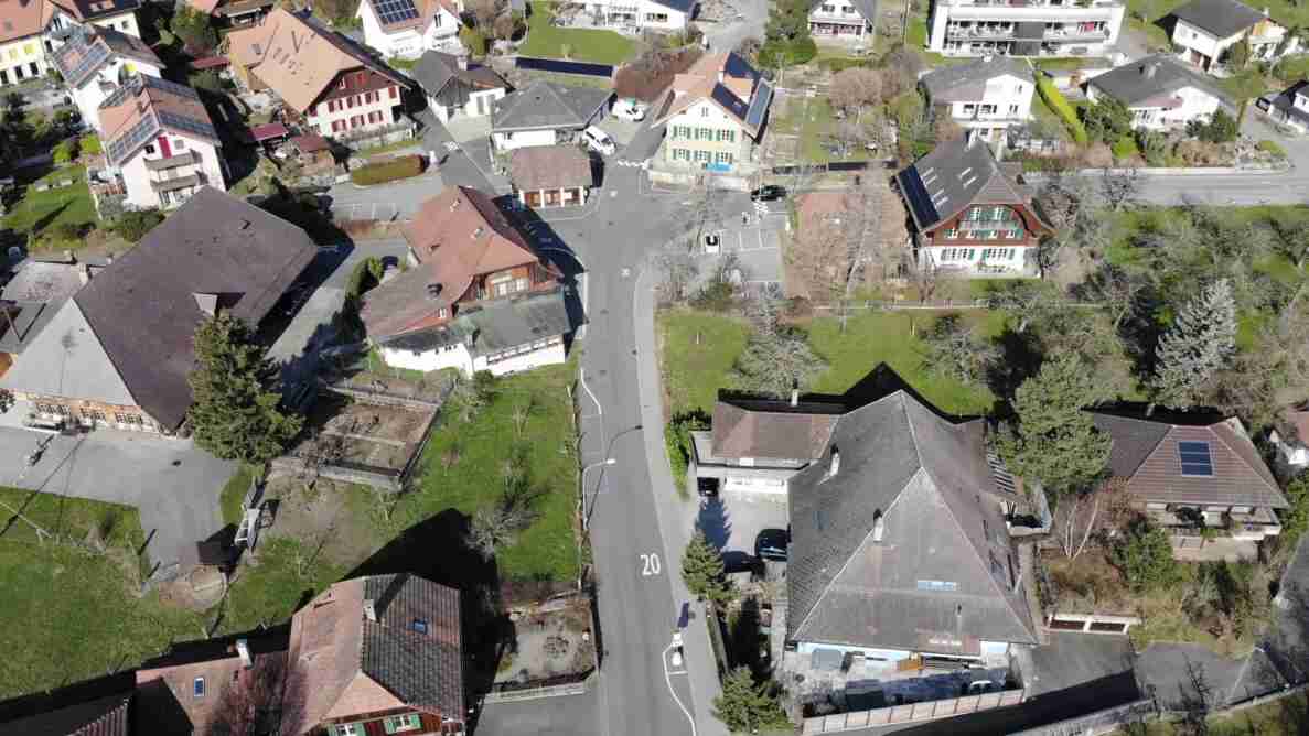 Luftaufnahme der Begegnungszone Habstetten mit auf die Fahrbahn aufgemalter Tempo-20-Markierung sowie einer 30er-Markierung im oberen Bildbereich. Die Strasse ist asphaltiert und in gutem Zustand, verläuft geschwungen durch das Dorf und ist von Einfamilienhäusern mit Ziegel- und Solardächern sowie Gärten gesäumt. Vereinzelt stehen parkierte Fahrzeuge am Strassenrand, eine Person ist auf einem Gehweg zu erkennen.