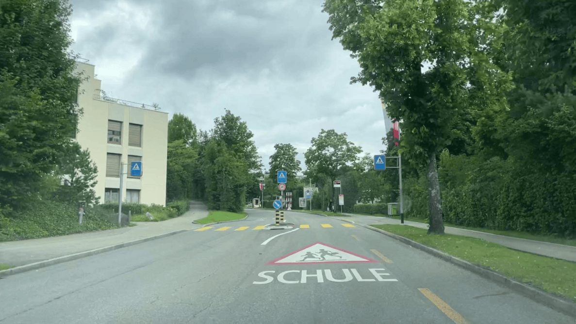 Strasse mit gelb markiertem Fussgängerstreifen und Fahrbahnmarkierung «SCHULE» mit Warndreieck für spielende Kinder. Blaue Fussgängerstreifen-Signale beidseitig sowie eine Tempo-40-Zone-Tafel und Verbotssignal für Fahrzeuge mit Anhänger sind sichtbar. Eine Verkehrsinsel mit gelb-schwarzer Markierung trennt die Fahrspuren. Die Strasse ist von Bäumen gesäumt, ein mehrstöckiges Gebäude steht links im Hintergrund.