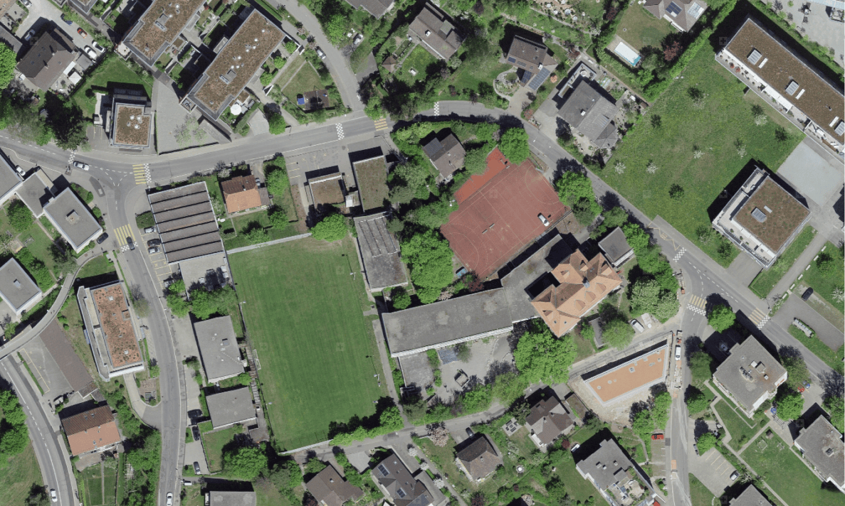 Senkrechtes Luftbild des Flugbrunnenareals in Bolligen mit Wohnhäusern, Rasenspielfeld, rotem Kunstrasenplatz und Schulgebäuden. Umliegende Quartierstrassen mit Fussgängerstreifen und Fahrbahnmarkierungen erschliessen das Areal, einzelne parkierte Fahrzeuge stehen am Strassenrand. Die Strassen sind in gutem Zustand, Personen sind keine erkennbar.