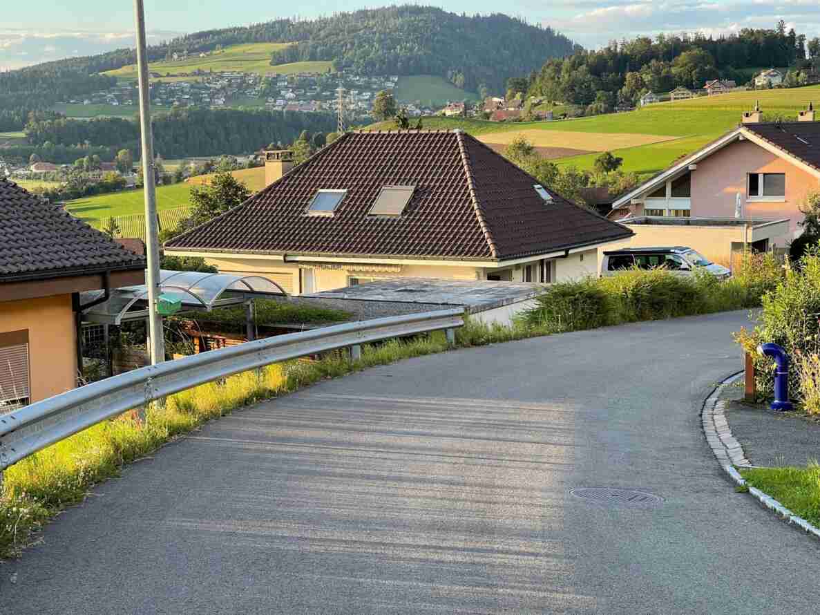 Kurvige, asphaltierte Quartierstrasse mit Leitplanke entlang eines Hanggrundstücks, ohne Verkehrssignale oder Verkehrsteilnehmende. Rechts ein blauer Hydrant, ein Kanaldeckel und ein Randstein markieren den Strassenrand. Blick über Einfamilienhäuser mit Ziegeldächern hinweg auf ein hügeliges Landschaftspanorama mit verstreuten Siedlungen, Wiesen und bewaldeten Hügeln im Hintergrund.