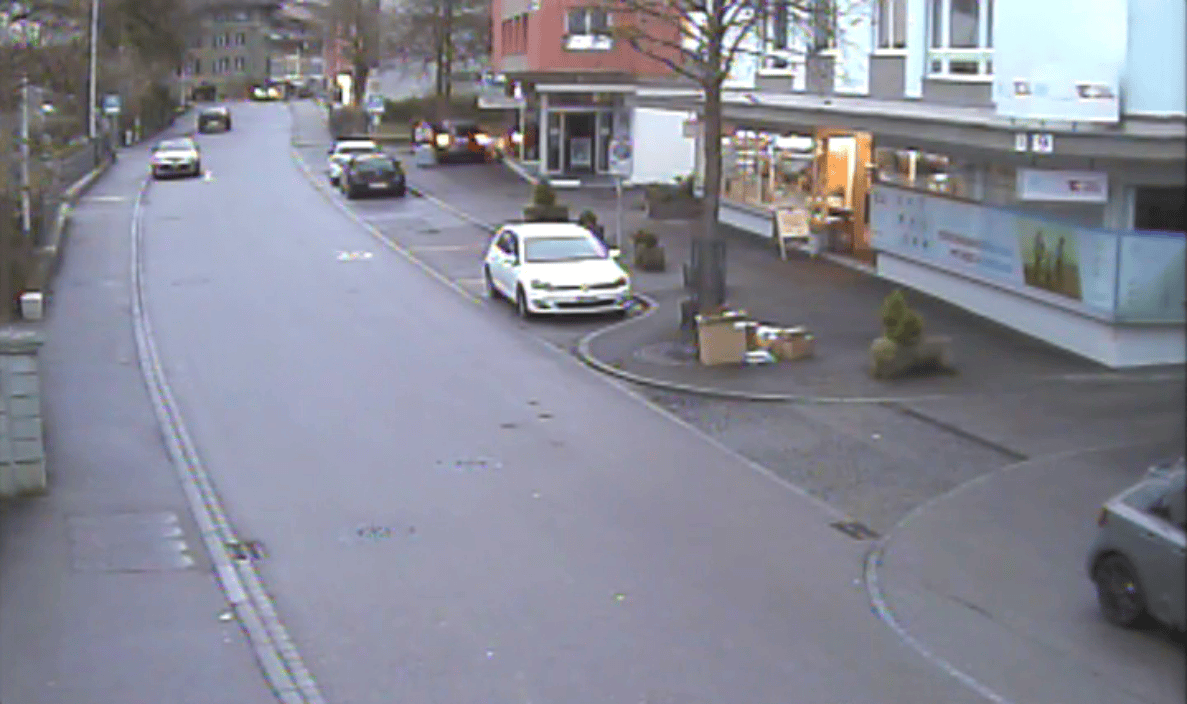 Videostandbild der Lyssachstrasse in Burgdorf mit Querungsbereich: mehrspurige Strasse mit parkierten und fahrenden Autos entlang von Geschäftshäusern, gepflästerter Vorplatz mit Bäumen und Pflanzkübeln vor einem Ladengeschäft, schmale Trottoirs beidseits der Fahrbahn, keine Fussgänger sichtbar, ruhiger Verkehr auf innerörtlicher Erschliessungsstrasse.