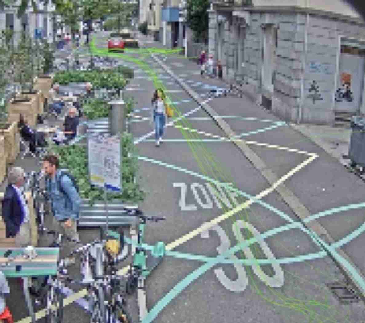Cramerstrasse mit Fahrbahnmarkierung «ZONE 30». Auf der Strasse sind farbige Linien zur Bewegungsanalyse von Fussgängerinnen, Velofahrenden und Fahrzeugen eingeblendet (Messung). Am Strassenrand stehen Sitzgelegenheiten, Pflanztröge und parkierte Velos, davor sitzen und stehen mehrere Personen. Eine Person überquert zu Fuss die Fahrbahn, im Hintergrund fährt ein rotes Fahrzeug. Die Strasse ist grosszügig für Fussverkehr und Veloverkehr gestaltet.