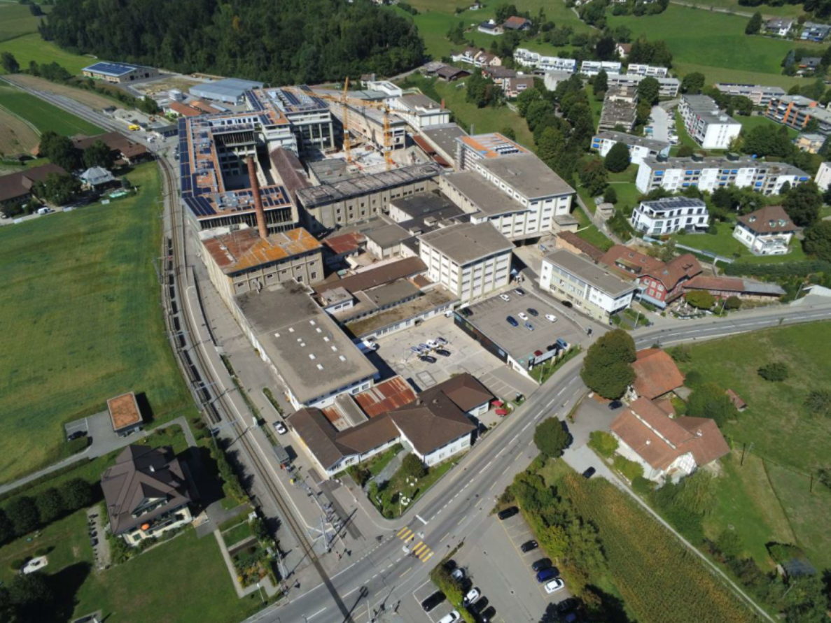 Drohnenaufnahme des Bernapark-Areals in Deisswil mit grossen Industrie- und Lagergebäuden, teils mit Solarpanels auf den Dächern und Baukränen im Hintergrund. Eine Bahnlinie verläuft entlang der Gebäude, daneben eine zweispurige Strasse mit Fussgängerstreifen und wenigen parkierten Fahrzeugen. Umliegend sind Wohnhäuser, Grünflächen und Bäume zu sehen, der Verkehr auf den Strassen ist gering.