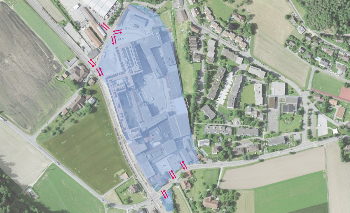 Luftbild von Deisswil bei Bernapark mit blau markiertem Areal des grossen Gebaeudekomplexes sowie umliegender Wohnsiedlung, Feldern und Bahnlinie. Pinke Doppelpfeile entlang der angrenzenden Strasse kennzeichnen mehrere Stellen mit Verkehrsfluss oder Messpunkten. Wohnstrassen mit Einfamilienhaeusern und Parkplaetzen umgeben das Areal, dazwischen liegen Gruenflaechen und landwirtschaftliche Parzellen.