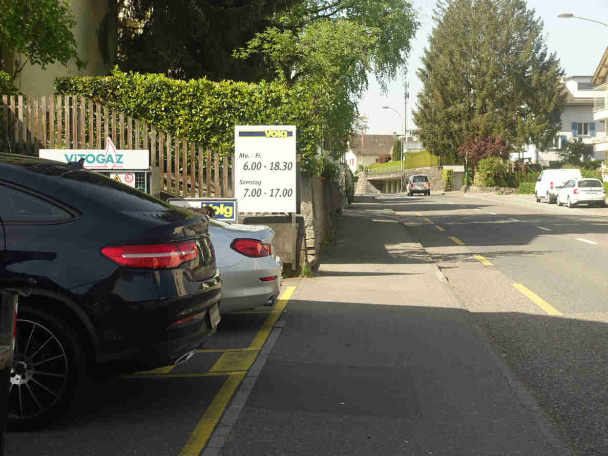 Strasse mit gelber Sicherheitslinie und Parkfeldern, in denen zwei Fahrzeuge parkiert sind. Rechts fährt ein Lieferwagen auf der Fahrbahn talwärts, weitere Autos stehen am Strassenrand. Im Hintergrund führt die Strasse leicht bergab zu einer kleinen Unterführung. Ein Hinweisschild zeigt die Öffnungszeiten eines Volg-Ladens, daneben ein Vitogaz-Schild an einem Holzzaun mit grüner Hecke.
