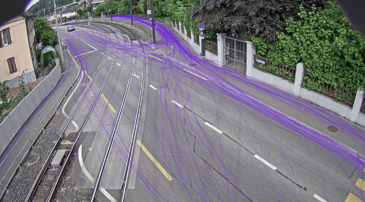 Überwachungskamerabild einer kurvigen Strasse mit Tramschienen, auf der violette Linien die Bewegungsverläufe von Fahrzeugen zur Verkehrsmessung markieren. Ein einzelnes Auto fährt im Hintergrund auf der Fahrbahn. Der Strassenbelag ist mit gelben und weissen Markierungen versehen, seitlich befinden sich Gebäude, Mauern und dichte Bepflanzung.