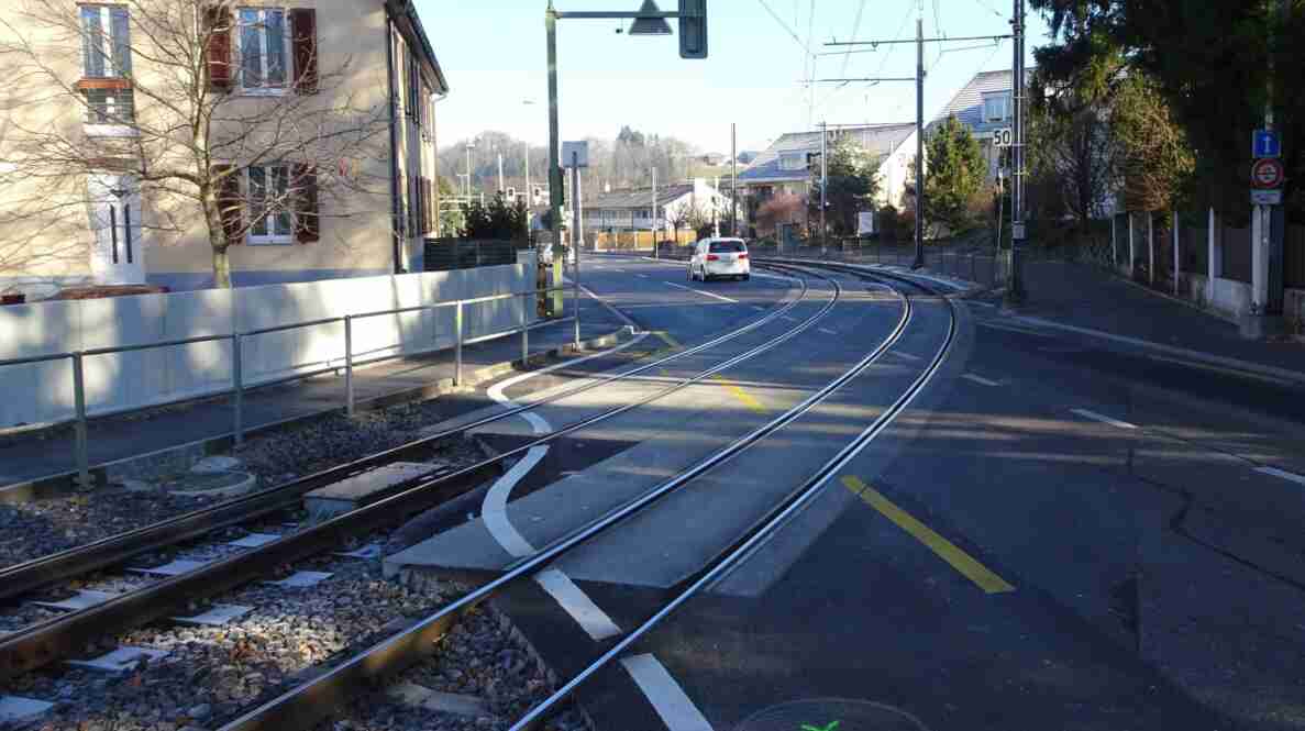 Bahnübergang Therwilerstrasse in Ettingen mit Gleisen der Bahnstrecke, die neben der Strasse verlaufen und in einer Kurve abzweigen. Die Fahrbahn ist trocken und frisch asphaltiert, mit gelben und weissen Markierungen im Bereich der Schienen. Ein Warnschild vor dem Bahnübergang sowie ein Tempo-50-Signal sind sichtbar. Ein weisses Auto fährt auf der Strasse Richtung Kreuzung, im Hintergrund Wohnhäuser und kahle Bäume.