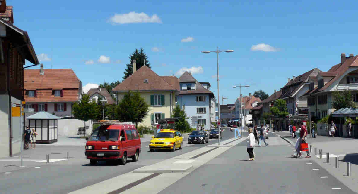 Verkehrsberuhigte Dorfstrasse mit begegnungszonenähnlicher Gestaltung, Mittelinsel und Pollern statt klassischer Trottoirs. Ein roter Toyota-Lieferwagen, ein gelbes Taxi sowie mehrere Personenwagen befahren die Strasse, ein Motorrad folgt. Mehrere Personen überqueren die Fahrbahn ausserhalb markierter Fussgängerstreifen, andere warten am Strassenrand. Umliegend stehen typische Schweizer Wohn- und Geschäftshäuser mit Ziegeldächern.