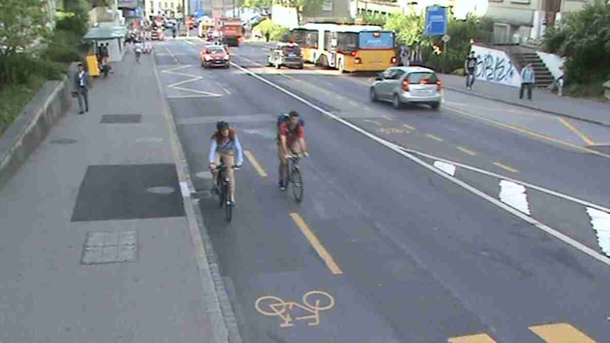 Blick von oben auf eine mehrspurige Innerortsstrasse mit Velo-Piktogramm auf der markierten Radspur. Zwei Radfahrende fahren hintereinander auf dem Radstreifen, während sich von hinten ein silberner Personenwagen sowie ein Linienbus nähern. Trottoirs mit Fussgängerinnen und Fussgängern säumen die trockene, gut sichtbar markierte Fahrbahn beidseitig.