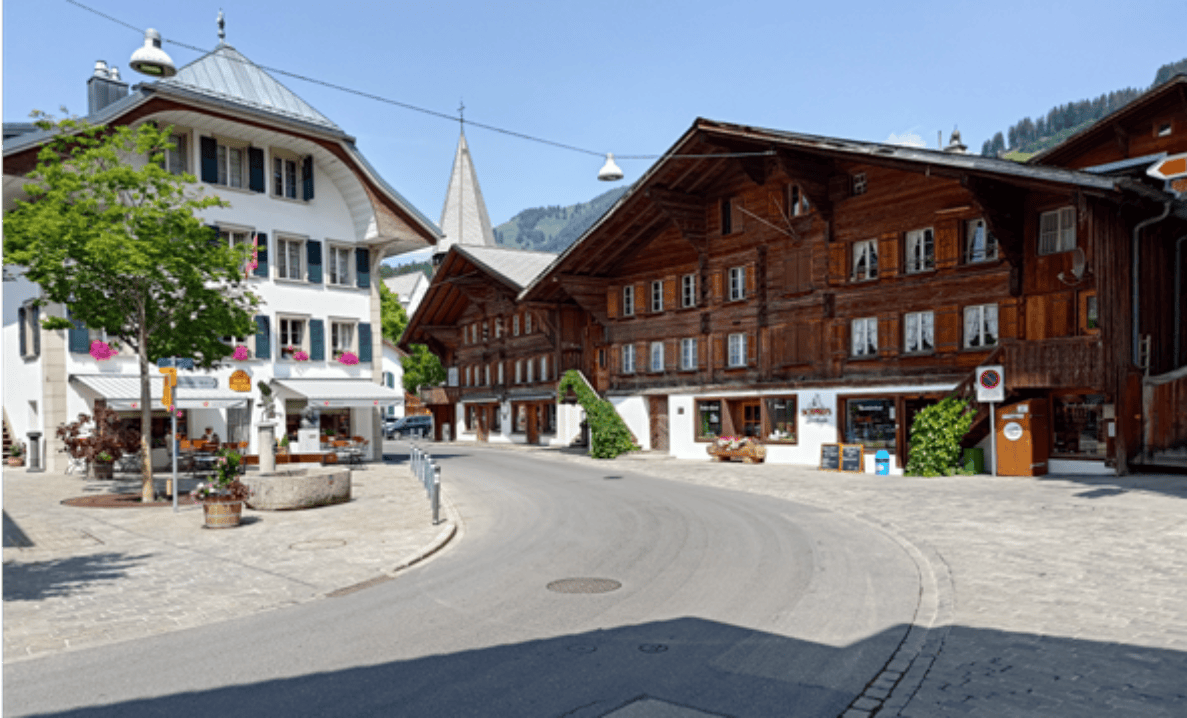 Gemeindestrasse im Ortskern von Saanen mit gepflästerter Kurve zwischen historischen Chalet-Gebäuden. Ein Fahrverbotsschild mit Zusatz steht rechts am Strassenrand, daneben ein Rundschild. Die Fahrbahn ist breit und autofrei, an einem Brunnen und Blumenschmuck vorbeiführend. Im Hintergrund ein Kirchturm, keine Personen auf der Strasse sichtbar.