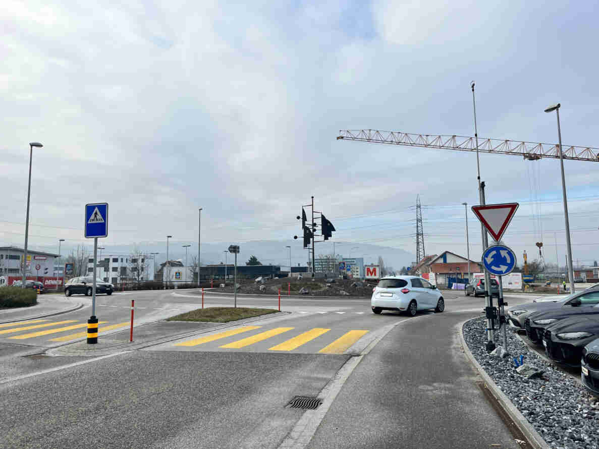 Kreisverkehr mit Vortrittssignal und Kreisel-Symbol, gelbem Fussgängerstreifen sowie Fussgängerstreifen-Hinweistafel. Ein weisses Auto durchfährt den Kreisel, weitere Fahrzeuge stehen parkiert. Auf einem Mast ist eine installierte Kamera zur Verkehrsmessung angebracht. Im Hintergrund Gewerbebauten mit Denner- und Aldi-Beschriftung, ein Baukran sowie eine Windmessanlage. Strassenbelag ist trocken und intakt.