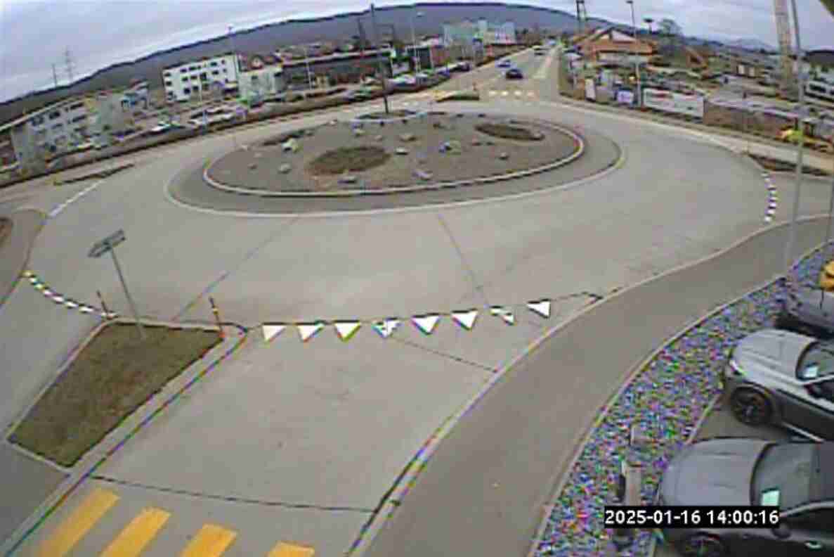 Videostandbild einer Verkehrsüberwachungskamera in Gretzenbach an der Oltnerstrasse im Grund. Zu sehen ist ein neu angelegter Kreisverkehr mit begrünter Mittelinsel, dessen Fahrbahn mit weiss-schwarzen Dreieckswimpeln provisorisch markiert ist. Ein Fussgängerstreifen führt zum Kreisel. Im Hintergrund fahren vereinzelt Fahrzeuge auf der Zufahrtsstrasse, im Vordergrund stehen parkierte Autos. Zeitstempel 2025-01-16, 14:00:16.