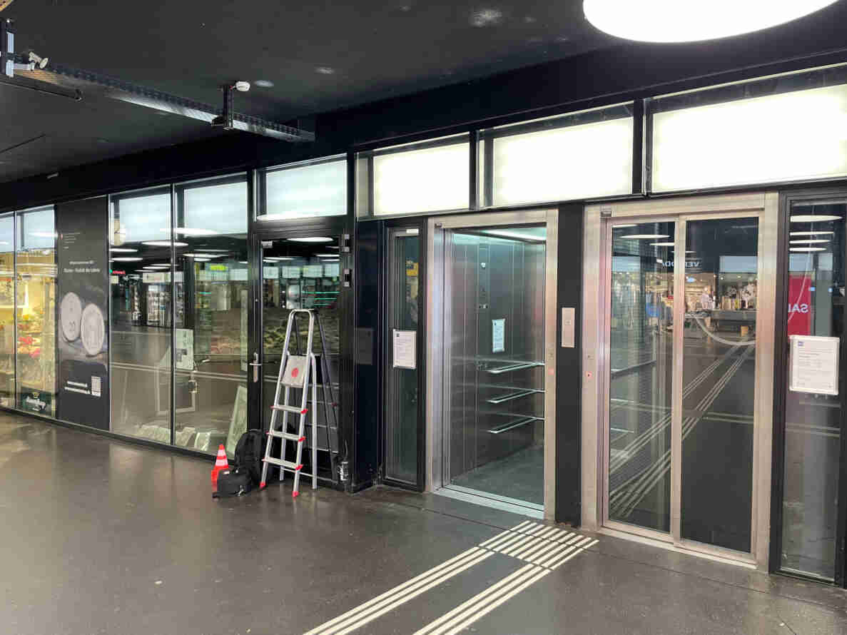 Innenbereich einer Passage mit Ladengeschäften, offener Liftkabine und danebenliegendem Aufzugsschacht mit Gitterschiebetüren. Vor dem Lift stehen eine Alu-Trittleiter, ein Rucksack und ein Verkehrshütchen. An der Decke ist eine Kamera zur Messung montiert. Am Boden verläuft ein taktiles Leitsystem für Sehbehinderte. Personen sind keine sichtbar.