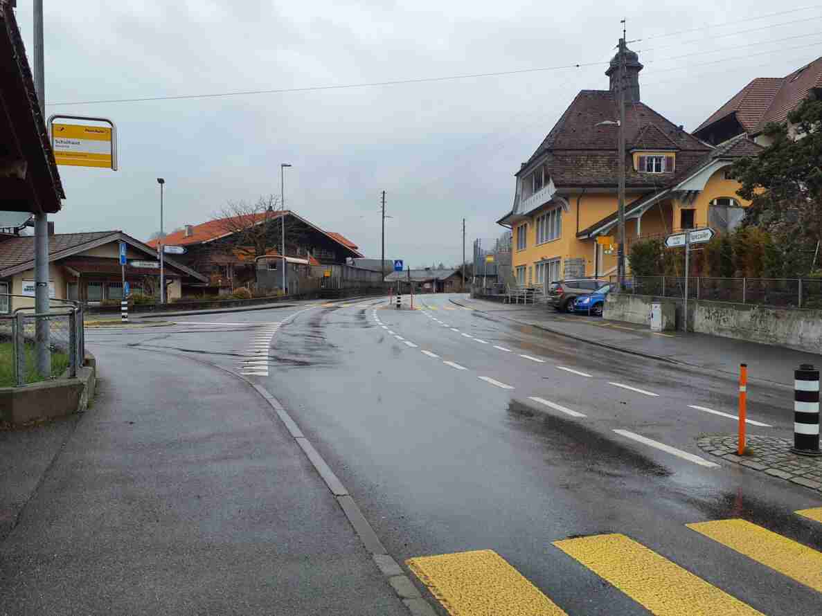 Gelber Fussgängerstreifen auf nasser Kantonsstrasse bei der Postauto-Haltestelle «Schulhaus Hondrich». Wegweiser zeigen Richtung Frutigen, Aeschi und Spiezwiler. Fahrbahnmarkierungen leiten den Verkehr um eine Verkehrsinsel mit rot-weiss markiertem Poller. Parkierte Autos stehen am Strassenrand vor gelben und braunen Gebäuden, keine Fussgänger sichtbar.
