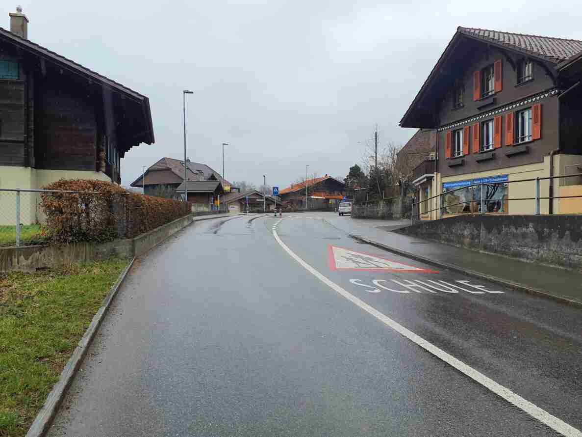 Ortsdurchfahrt Hondrich in Blickrichtung Süd mit nasser Fahrbahn. Auf dem Asphalt ist eine Fahrbahnmarkierung mit Schulwarnsymbol und Schriftzug «Schule» aufgemalt, ergänzt durch ein blaues Fussgängerstreifen-Signal am Strassenrand. Die kurvig verlaufende Strasse mit Leitlinien führt durch das Dorf vorbei an typischen Berner Chalets, ein einzelnes Auto fährt entgegenkommend. Keine Personen sind auf der Strasse sichtbar.