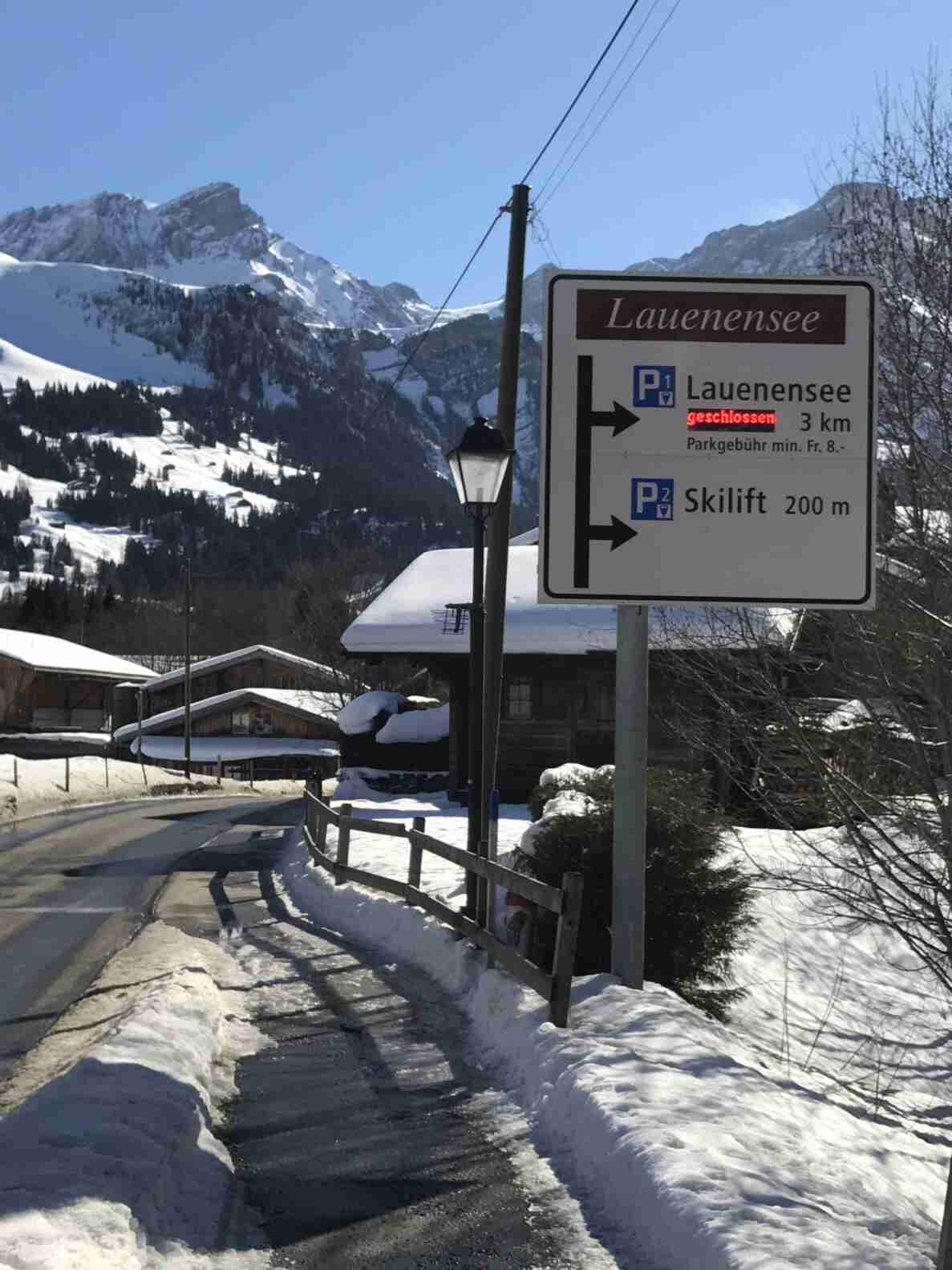 Wegweiser «Lauenensee» am Strassenrand zeigt Parkleitsystem mit P1 Lauenensee (3 km, aktuell «geschlossen» als LED-Anzeige, Parkgebühr min. Fr. 8.-) sowie P2 Skilift (200 m), beide nach rechts weisend. Die schneegeräumte, teils vereiste Nebenstrasse führt an verschneiten Holzchalets vorbei, im Hintergrund schneebedeckte Berggipfel. Keine Fahrzeuge oder Personen sichtbar.