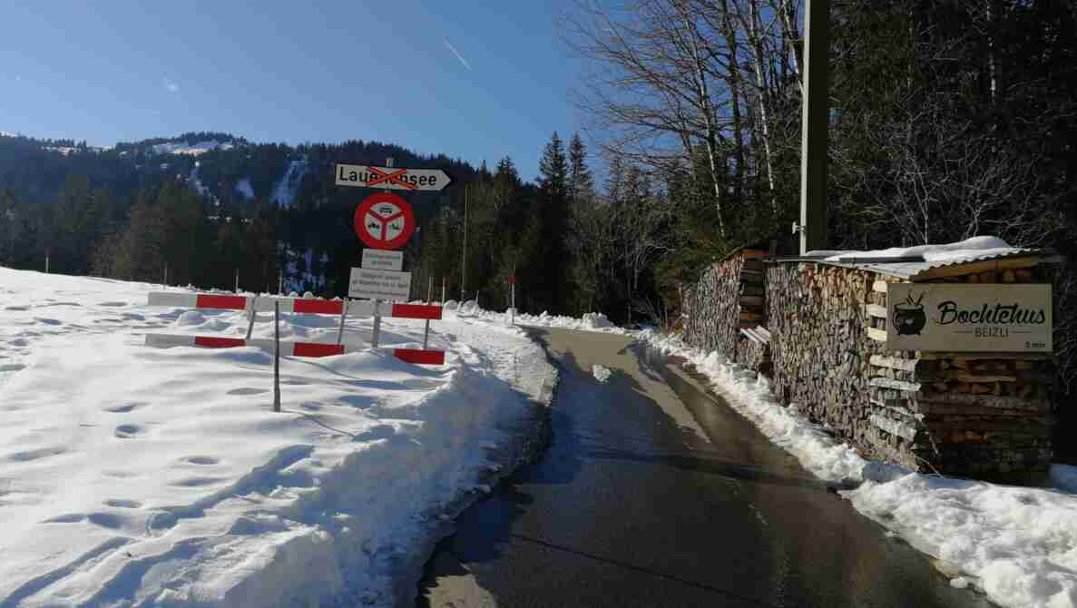 Strasse zum Lauenensee mit nasser, schneegeräumter Fahrbahn und schneebedeckten Rändern. Ein Wegweiser Lauenensee ist mit rotem Kreuz durchgestrichen, darunter ein Verbotssignal für Motorwagen und Motorräder mit Zusatztafel «Zubringerdienst gestattet» und Gültigkeit November bis April. Eine rot-weisse Barriere sperrt die Strasse zusätzlich. Rechts ein Holzstapel und ein Wegweiser zum «Bochtehus Beizli», 5 Minuten entfernt.