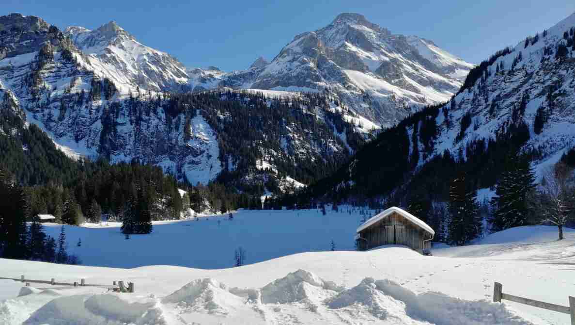 Blick auf den schneebedeckten Lauenensee, umgeben von schneebedeckten Berggipfeln und Nadelwäldern. Am Ufer steht eine kleine Holzhütte mit schneebedecktem Dach, im Vordergrund ein Holzzaun und tiefe Schneeverwehungen mit Fussspuren. Es sind keine Verkehrssignale, Strassen, Fahrzeuge oder Messgeräte sichtbar.
