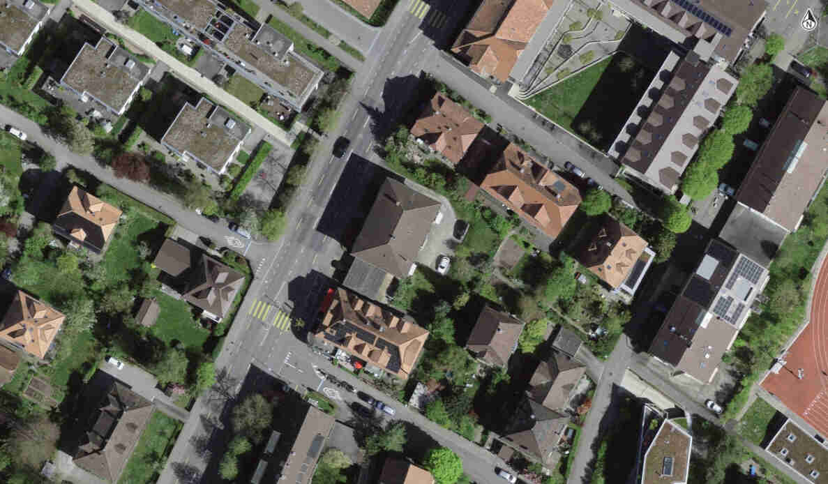Senkrechtes Luftbild eines Wohnquartiers mit Einfamilienhäusern, Steildächern und Gärten. Eine Hauptstrasse verläuft diagonal durchs Bild mit Sperrflächen-Markierungen, Fussgängerstreifen und einer kleinen Kreuzung im Zentrum. Parkierte Autos stehen entlang der Strassen, einzelne Fahrzeuge sind in Bewegung. Rechts unten ist eine Laufbahn eines Sportplatzes sichtbar. Grundlage @swisstopo.