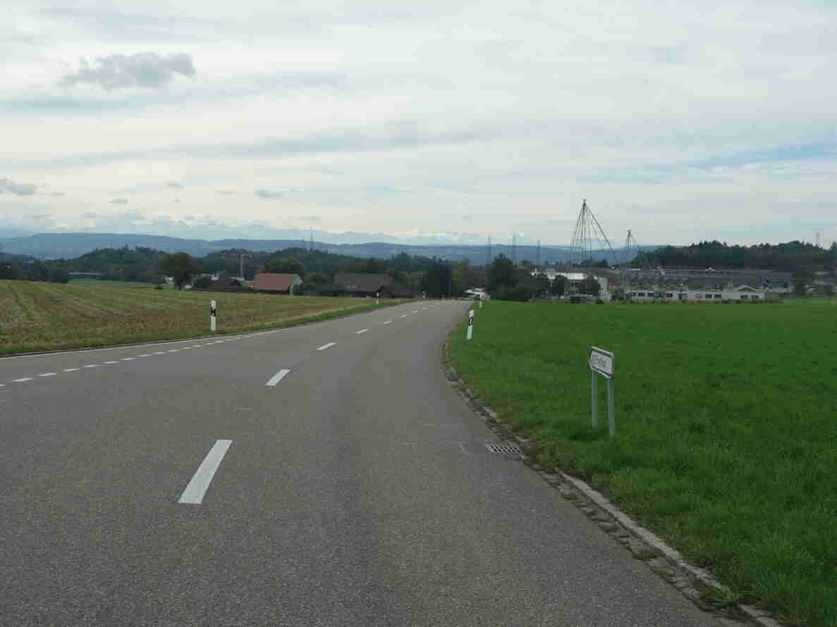 Ausserortsabschnitt der Brüttenerstrasse in Blickrichtung Süden, eine zweispurige Landstrasse durch offenes Landwirtschaftsgebiet. Fahrbahn mit Mittelmarkierung und Randleitpfosten, gut befahrbarer Asphaltbelag. Rechts ein Wegweiser mit Ortsbezeichnung Effretikon. Im Hintergrund ein Ortsteil mit Gebäuden, Hochspannungsmasten und Alpenpanorama. Keine Fahrzeuge oder Personen im Vordergrund erkennbar.