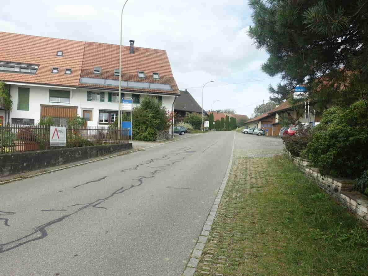 Brüttenerstrasse in der Ortschaft Kleinikon mit Bushaltestellen-Schild, gesäumt von Bauernhäusern mit Ziegeldächern. Die asphaltierte Strasse weist geflickte Risse auf, rechts verläuft ein Pflastersteinstreifen entlang der Grasfläche. Am Strassenrand parkierte Autos, im Hintergrund gehen zwei Personen auf einem gepflasterten Platz. Bedeckter Himmel über der leicht ansteigenden Strasse.
