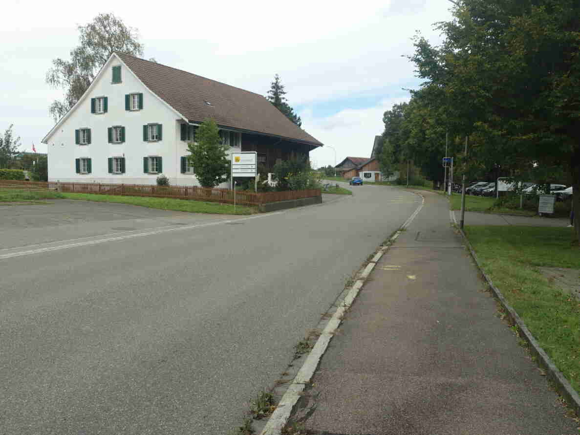 Brüttenerstrasse in Eschikon mit Blick auf ein Bauernhaus des Strickhofs, davor ein Wegweiser zu Gärtnerei und Hofladen. Die Strasse ist asphaltiert und ohne Mittellinie, ein separater Gehweg verläuft rechts entlang der Baumreihe. Ein blaues Auto fährt auf der Strasse, im Hintergrund parkierte Fahrzeuge auf einem Parkplatz. Eine Person ist am rechten Bildrand erkennbar.