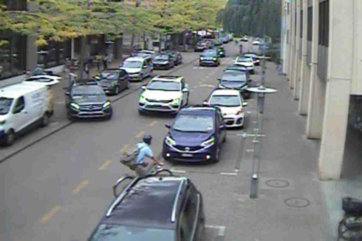 Bahnhofstrasse in Lyss mit dichtem Kolonnenverkehr in beide Richtungen. Mehrere Personenwagen stehen oder fahren langsam nebeneinander, dazwischen ein Lieferwagen. Auf einem separaten Trottoir mit Pollern gehen Fussgänger, eine Person mit Rucksack überquert die Fahrbahn zwischen den Fahrzeugen. Am rechten Bildrand ist ein Velo sichtbar. Die Strasse ist beidseitig von Gebäuden gesäumt.