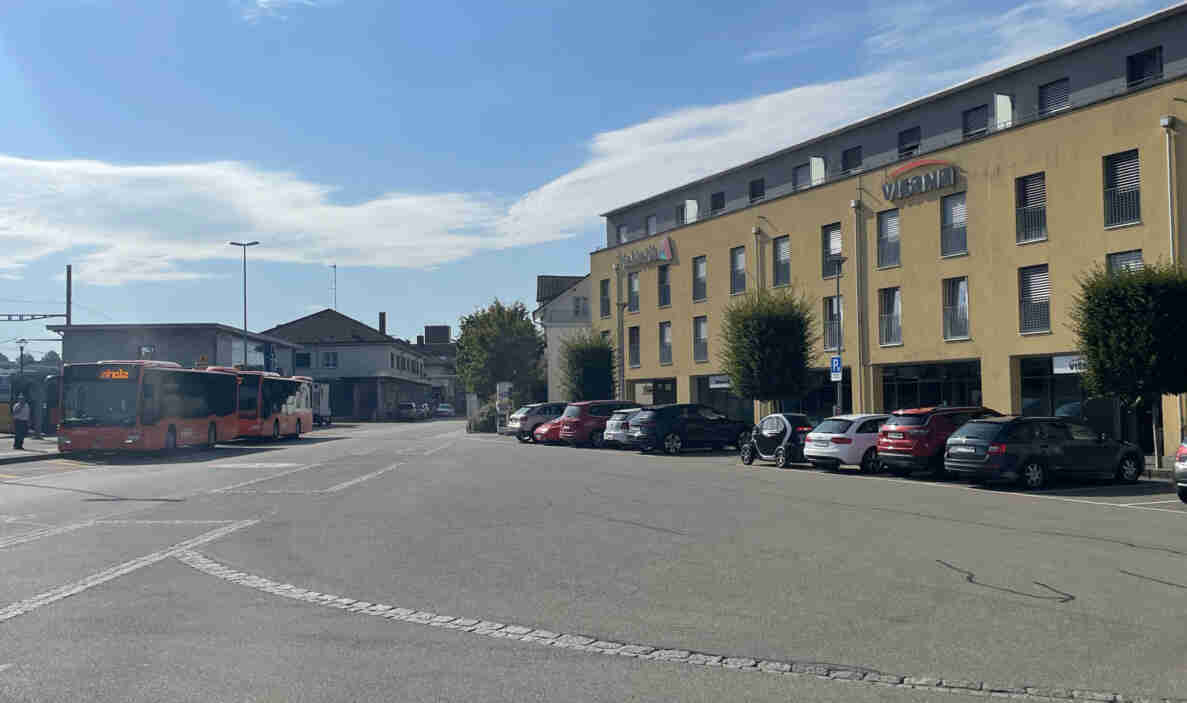 Bahnhofsplatz mit Bushaltestelle, an der zwei rote Gelenkbusse warten, im Hintergrund eine wartende Person. Rechts asphaltierte Fläche mit Parkfeldern, auf denen mehrere Personenwagen sowie ein Elektro-Kleinstfahrzeug stehen. Ein blaues Parkschild markiert einen Bereich mit eingeschränkter Parkzeit. Die Strasse ist gut instand gehalten und dient als Zubringer zum Busterminal. Im Hintergrund ein mehrstöckiges Gebäude mit den Schriftzügen «Visana» und «helvetia».