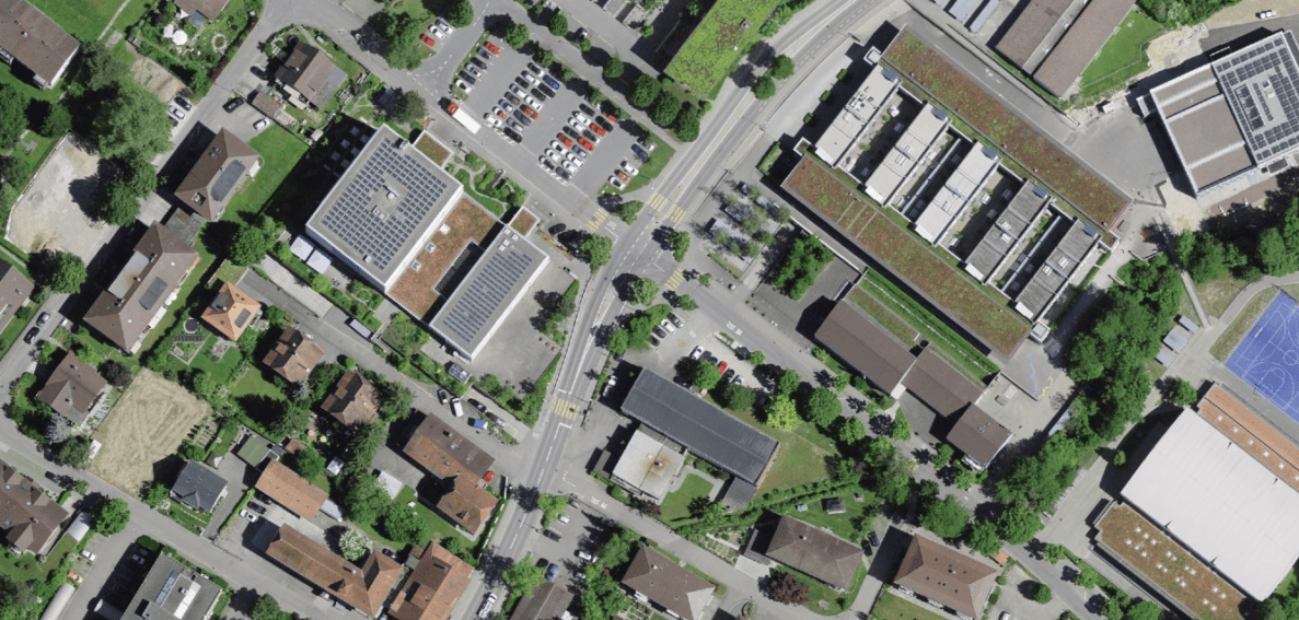 Senkrechte Satellitenaufnahme eines Wohnquartiers mit Kreuzung zweier Strassen, Fussgängerstreifen und gelben Fahrbahnmarkierungen im Kreuzungsbereich. Umliegend Einfamilienhäuser mit Gärten, grössere Mehrfamilienhäuser mit begrünten Dächern und Solaranlagen, Parkplätze mit mehreren abgestellten Fahrzeugen sowie ein Sportplatz. Strassen sind asphaltiert und in gutem Zustand, keine Fahrzeuge im Kreuzungsbereich sichtbar.
