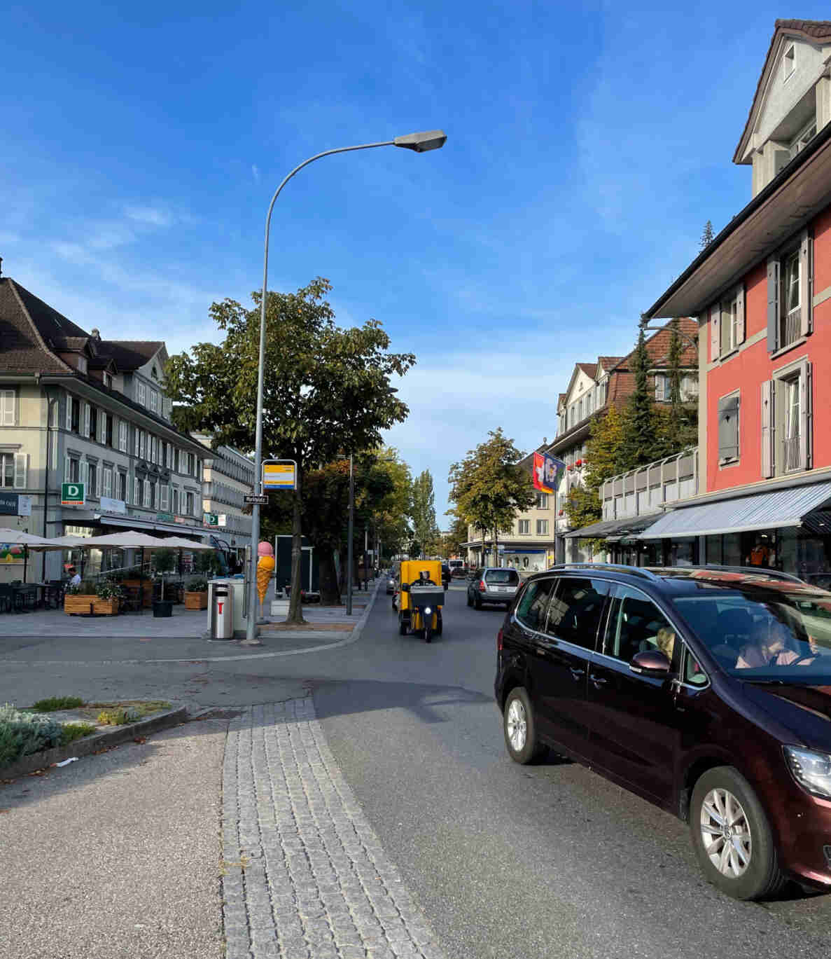 Strassenkreuzung beim Marktplatz mit Bushaltestellen-Schild, gepflästerter Fussgängerinsel und Fahrbahn in gutem Zustand. Ein dunkler Van fährt heran, dahinter ein gelbes Post-Lieferdreirad und weitere Autos folgen der Strasse zwischen historischen Gebäuden mit Ladengeschäften und Strassencafés. Bäume und ein Lichtmast säumen den Platz, im Hintergrund weht eine Kantonsflagge an einer rötlichen Fassade.