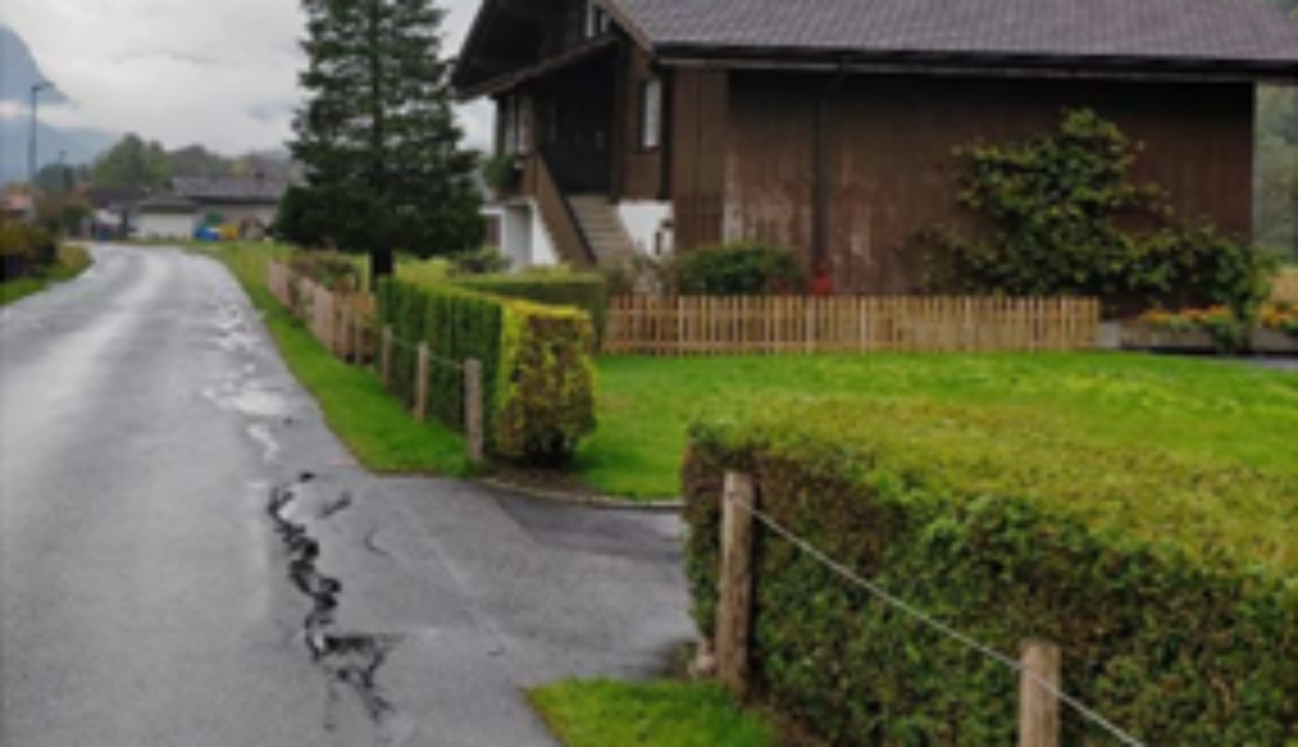 Schmale Quartierstrasse an der Balmstrasse in Meiringen, gesäumt von Hecken, Holzzaun und Bäumen. Eine private Ausfahrt zwischen den Hecken bietet eingeschränkte Sichtweite auf die Strasse, ohne Trottoir oder Randmarkierung. Die Fahrbahn weist Risse im Belag auf. Weder Fahrzeuge noch Personen sind auf der Strasse sichtbar.