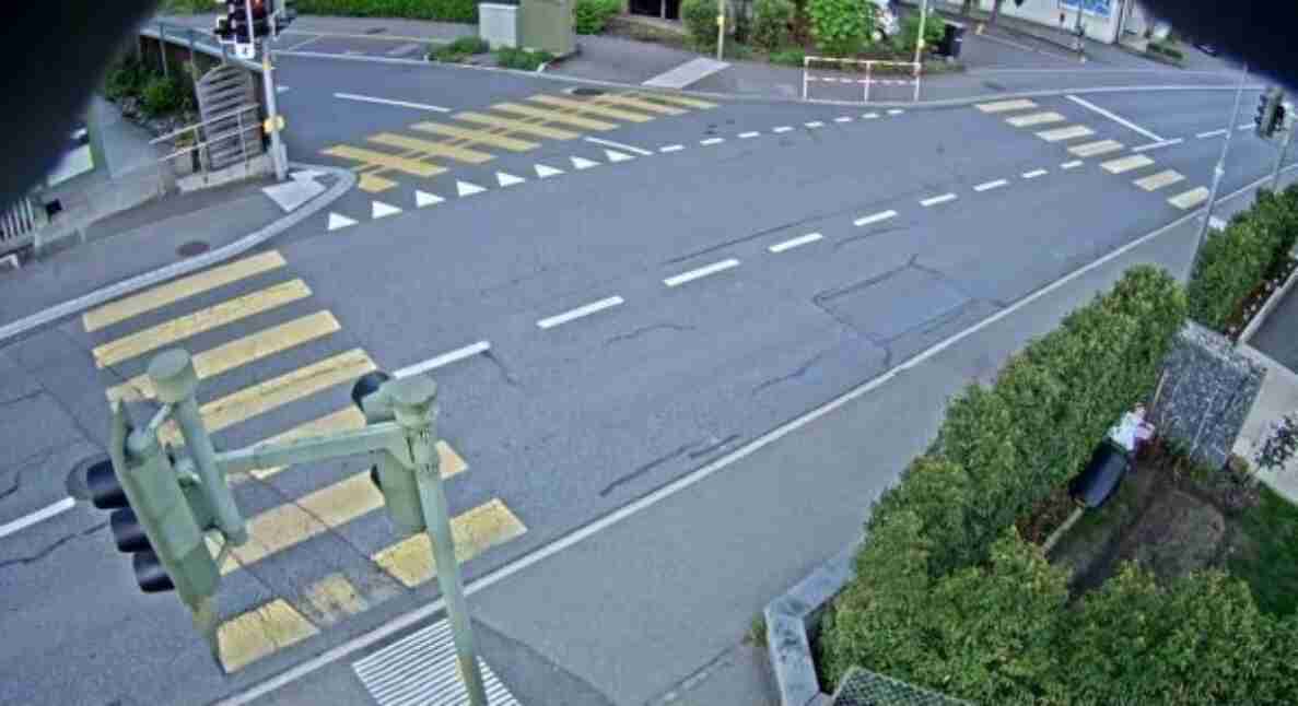 Videostandbild einer Verkehrskamera am Knoten Taegertschistrasse/Krankenhausweg in Muensingen. Übersicht auf eine breite Strassenkreuzung mit mehreren gelb-weissen Fussgaengerstreifen und Haltelinien. Im Vordergrund eine Ampelanlage mit Fussgaenger-Signal, im Hintergrund eine Bahnschranke sowie weitere Lichtsignale. Die Fahrbahn ist leer, es sind keine Fahrzeuge oder Personen unterwegs.