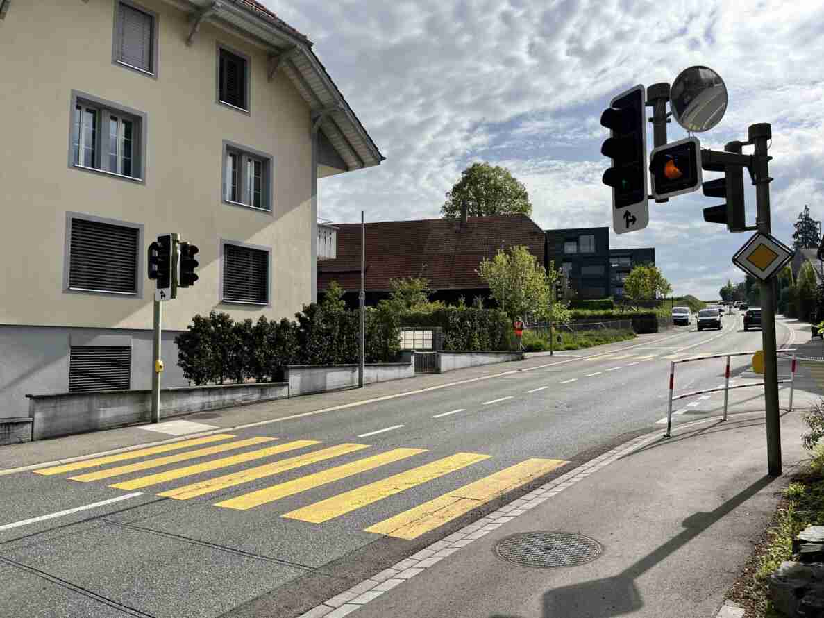 LSA-Knoten Tägertschistrasse/Krankenhausweg: Kreuzung mit Lichtsignalanlage, die Rot- und Orangephasen sowie Richtungspfeile für Linksabbiegen und Geradeausfahrt zeigt. Ein gelber Fussgängerstreifen führt über die Fahrbahn, ergänzt durch einen Vorsignal-Verkehrsspiegel und eine Baustellenabschrankung am Strassenrand. Der Asphalt ist intakt und trocken, im Hintergrund nähern sich mehrere Fahrzeuge sowie eine Person in oranger Warnweste, die sich dem Gehweg nähert.