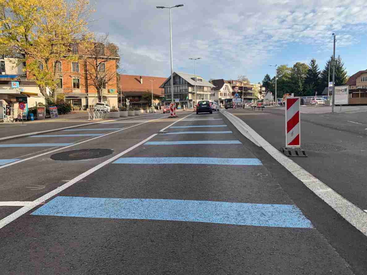Strasse mit frisch aufgemalten blauen Markierungen, vermutlich für eine provisorische Fahrbahnführung, und einer Verkehrsinsel mit rot-weisser Absperrbake. Verkehrsschilder zeigen ein Halteverbot sowie ein Kreisverkehr-Signal mit Wegweisern. Mehrere Personenwagen und ein Lieferwagen befinden sich im ruhig fliessenden Verkehr. Umliegend Ladengeschäfte, Wohnhäuser und Bäume mit Herbstlaub säumen die Strasse.